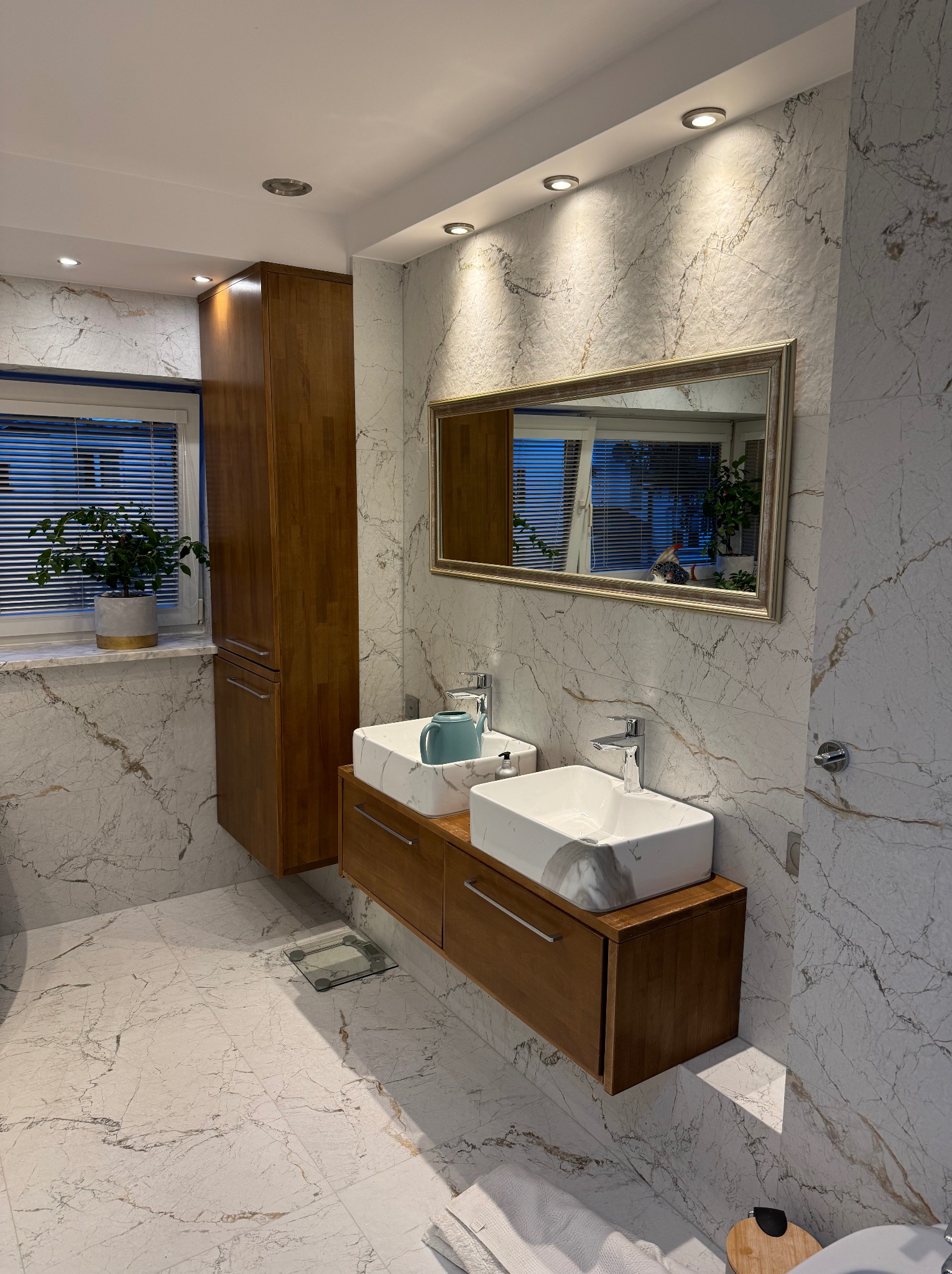 Nowoczesna łazienka z marmurowymi płytkami, drewnianą szafką pod umywalką i dużym lustrem. Umywalki nablatowe w kształcie prostokąta.