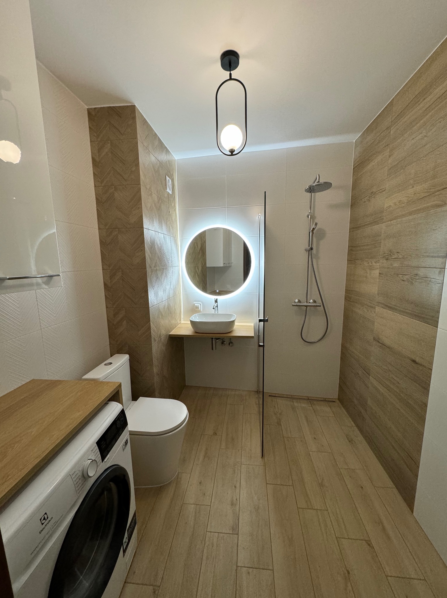 Nowoczesna łazienka z drewnopodobnymi płytkami, okrągłym lustrem z podświetleniem LED, pralką i kabiną prysznicową typu walk-in. Minimalistyczny design i ciepła kolorystyka.