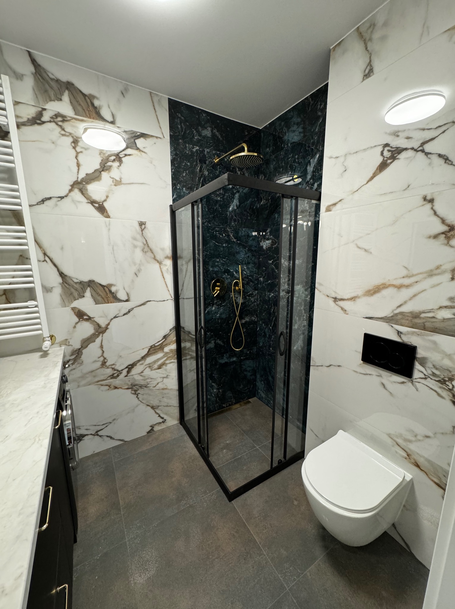 Nowoczesna łazienka z kabiną prysznicową typu walk-in, marmurowymi płytkami na ścianach i szarą podłogą. Armatura w kolorze złotym dodaje elegancji.