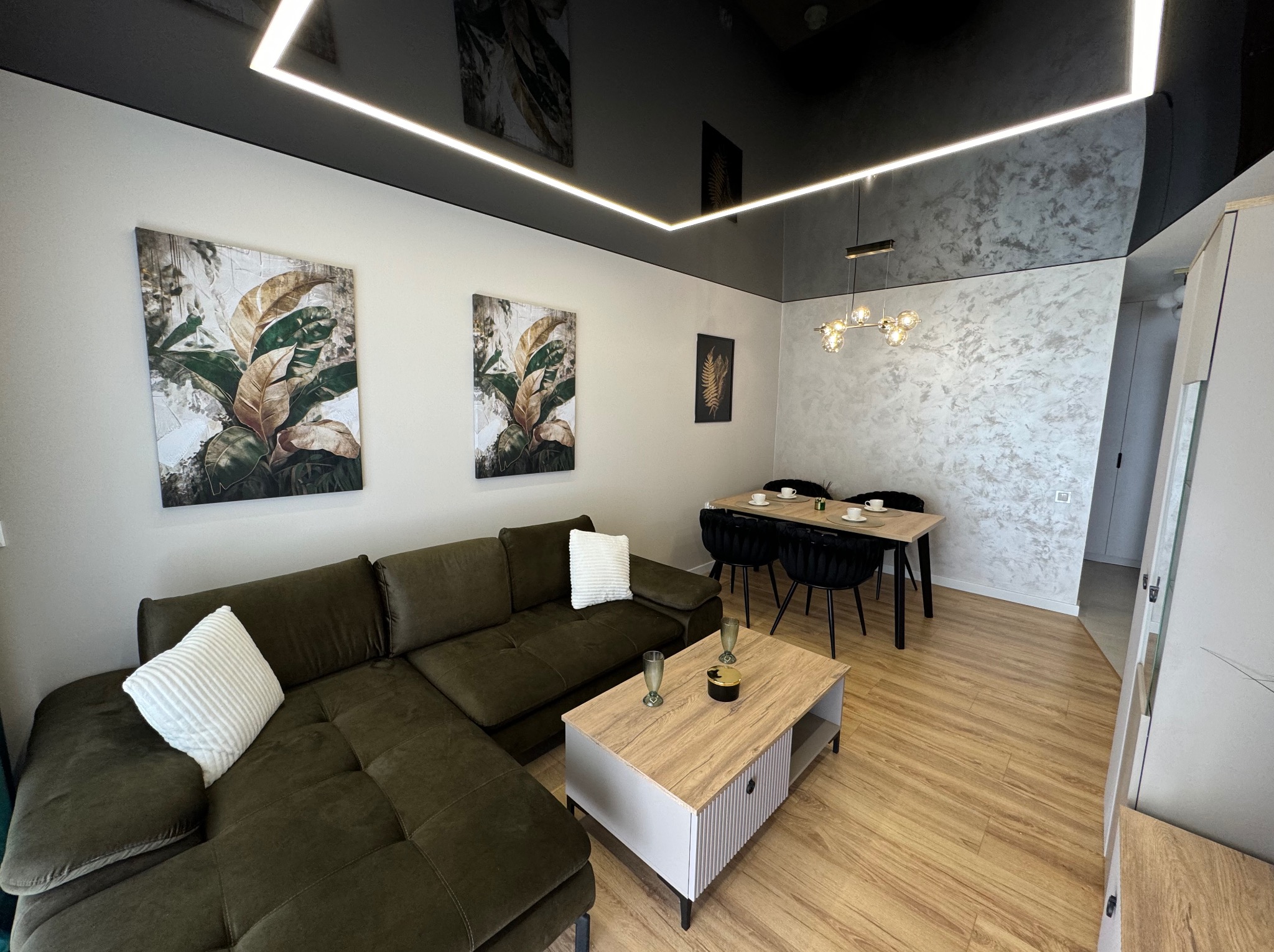 Stylowe wnętrze salonu z narożnikiem w odcieniach zieleni, drewnianym stolikiem kawowym i jadalnią. Na ścianach obrazy z motywami roślinnymi i dekoracyjny tynk.