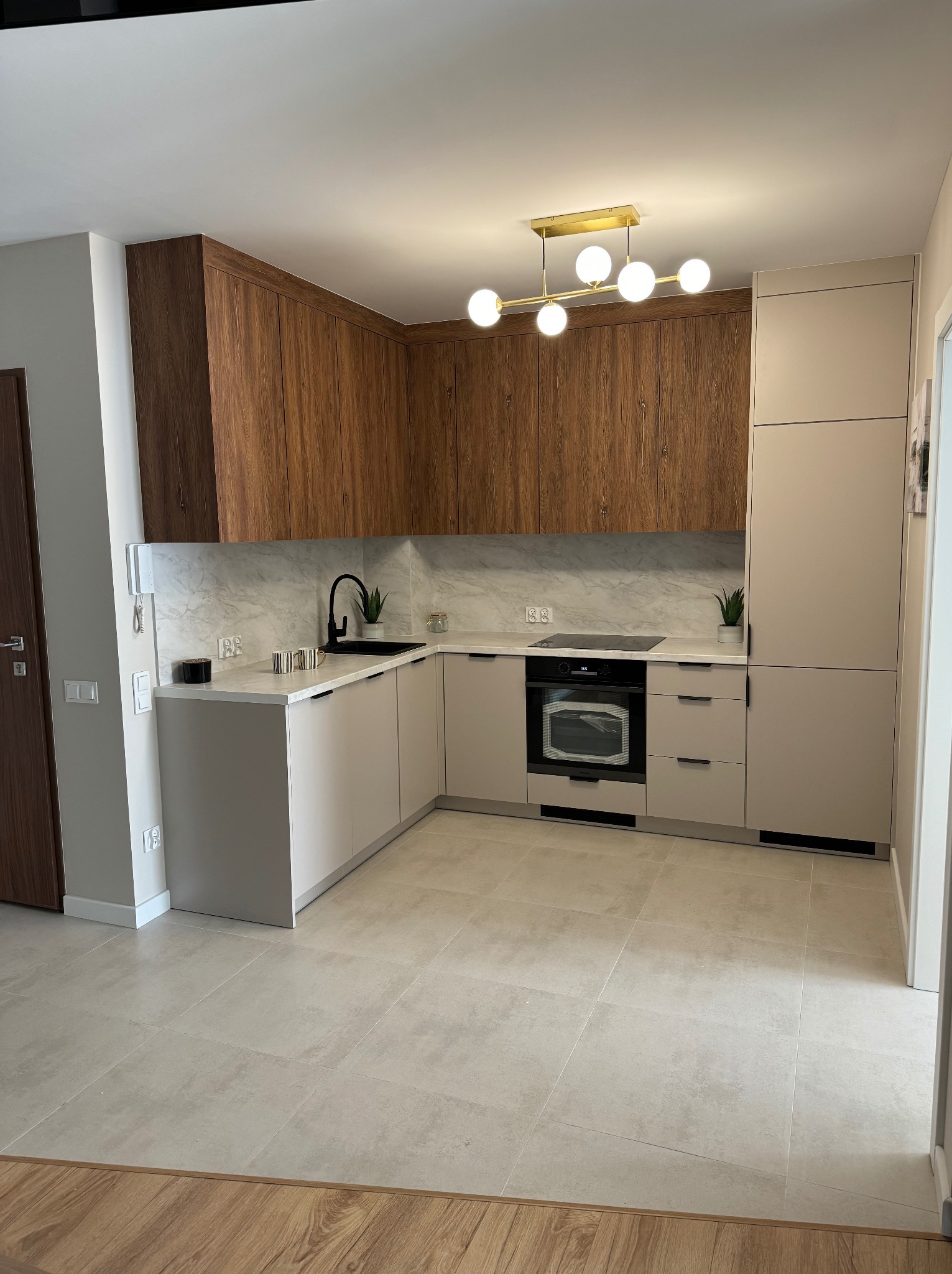 Nowoczesna kuchnia w beżach i brązach z marmurowym blatem, oświetlona złotą lampą sufitową. Uwagę zwraca czarna armatura i minimalistyczne dodatki.