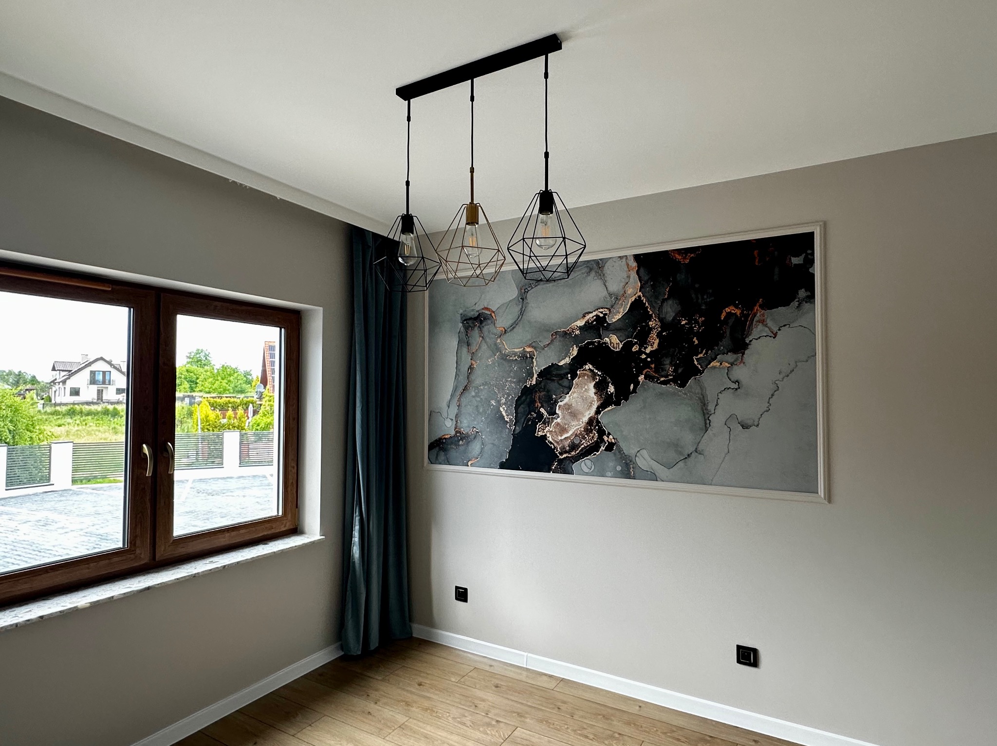 Wykończone wnętrze pokoju w Tarnowie: drewniana podłoga, okno z widokiem na zieleń, designerska lampa wisząca i obraz w marmurowym stylu na ścianie.