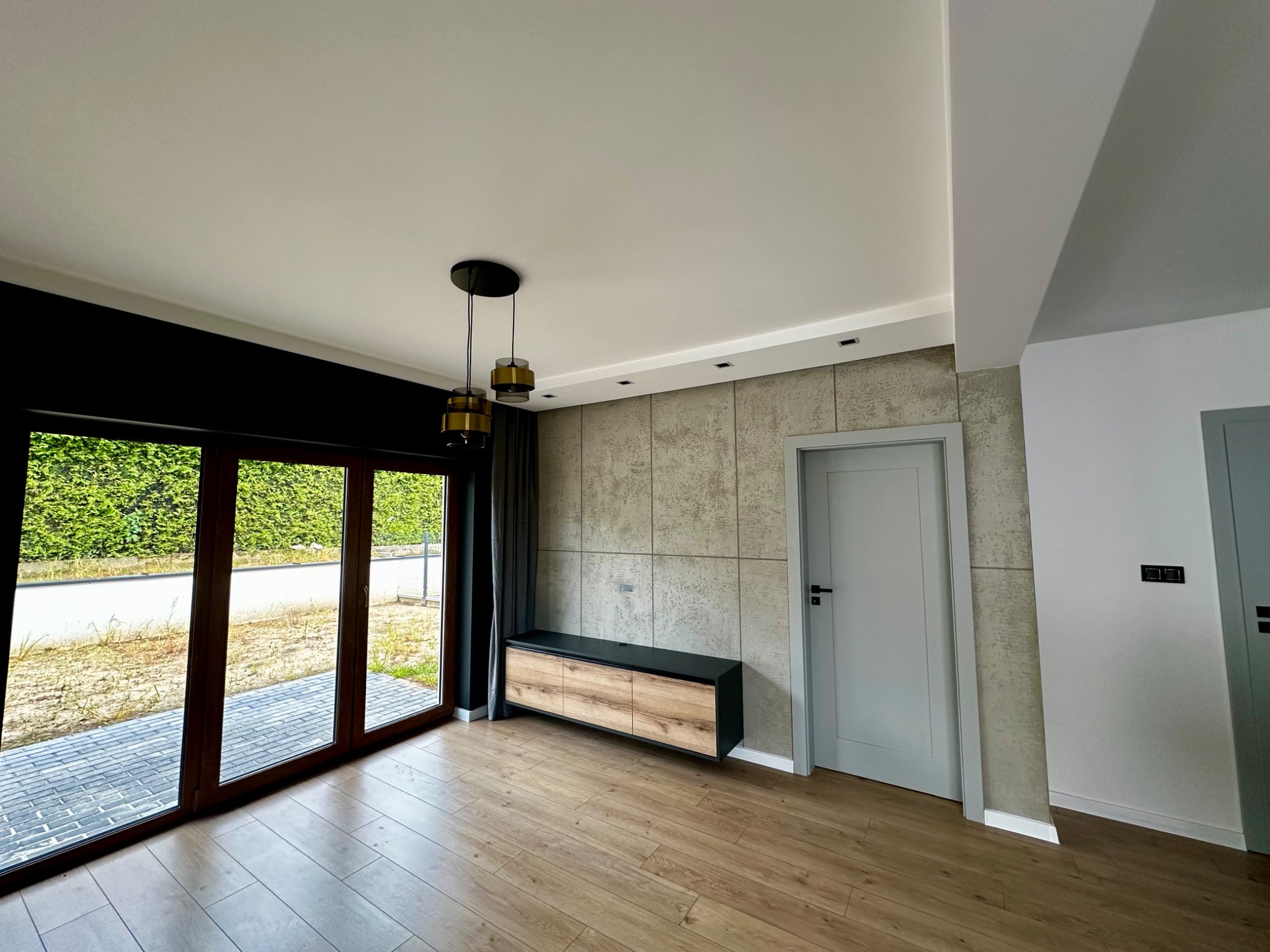 Nowoczesne wnętrze po remoncie: drewniana podłoga, ściana z betonową fakturą, szafka RTV, okno z widokiem na zieleń. Minimalistyczny design, jasne kolory.