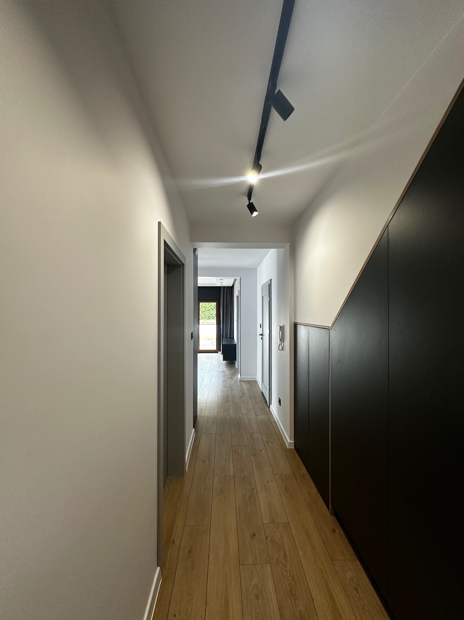 Nowoczesny korytarz z drewnianą podłogą, białymi ścianami i czarną zabudową. Oświetlenie punktowe na suficie. Widoczne drzwi i fragment wnętrza w głębi.