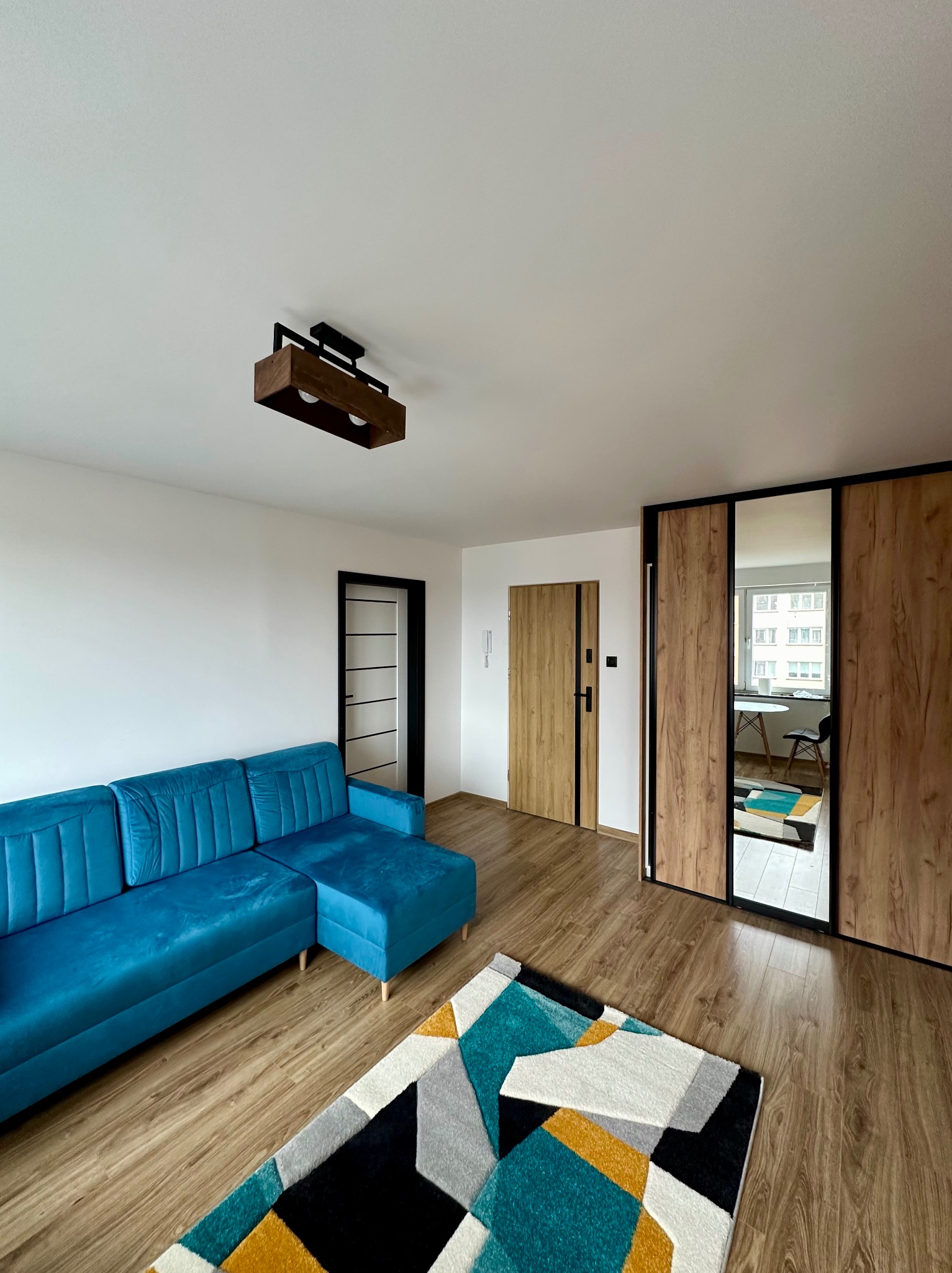 Jasne wnętrze po remoncie: turkusowy narożnik, drewniana podłoga, szafa z lustrami i drzwi w czarnych ramach. Nowoczesna lampa sufitowa.