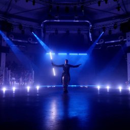 Jerzy Kozłowski - Oświetlona scena z osobą prezentującą lightshow z użyciem świetlnych rekwizytów, widzowie w tle, niebieskie światło, widowisko.