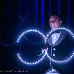 Jerzy Kozłowski - Mężczyzna w smokingu prezentuje lightshow z użyciem rekwizytów LED, tworząc świetlne wzory na ciemnym tle. Widoczne niebieskie pionowe pasy światła.