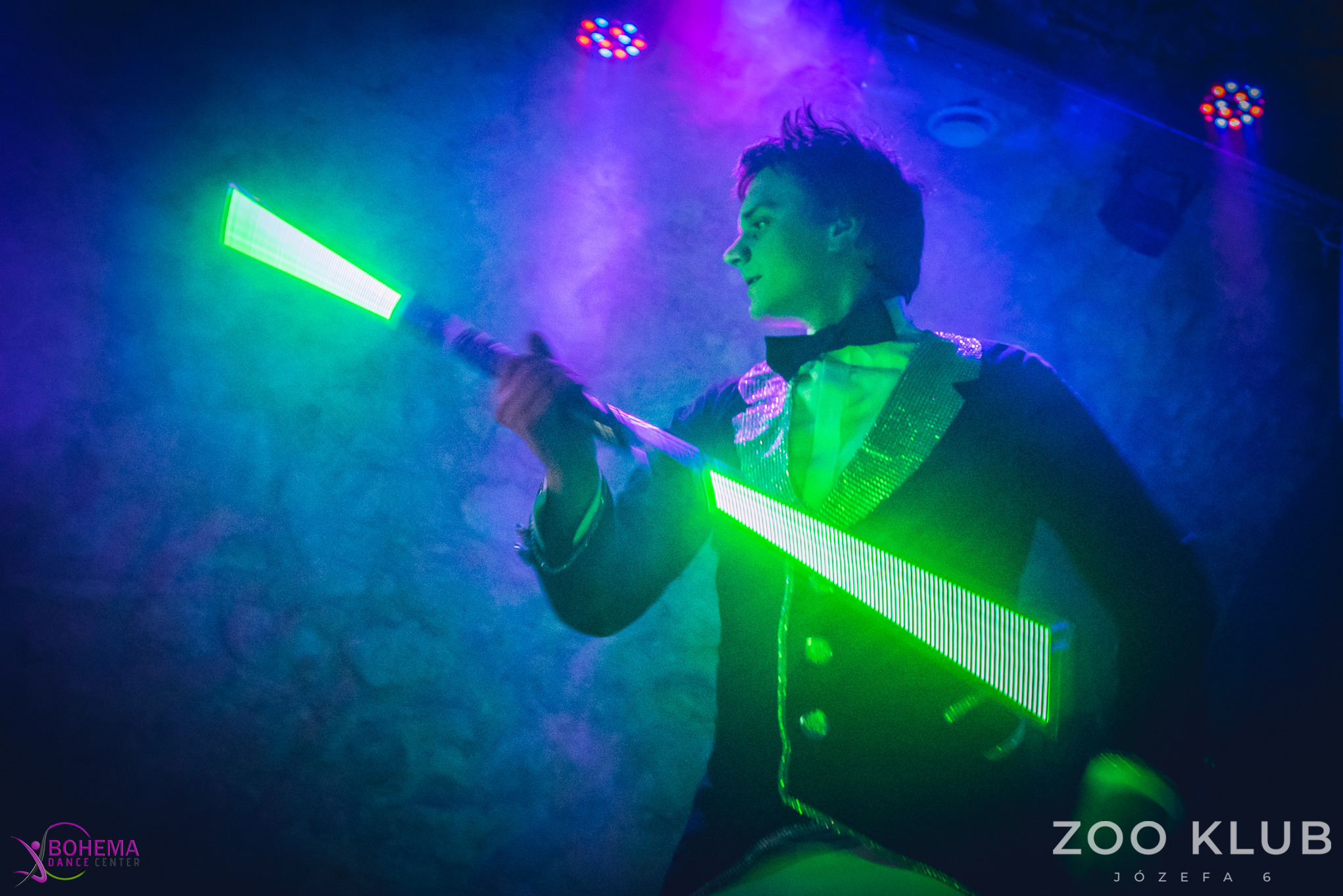 Mężczyzna w cekinowej marynarce trzymający świecące, zielone rekwizyty na tle klubu z oświetleniem scenicznym. Lightshow w Zoo Klub.