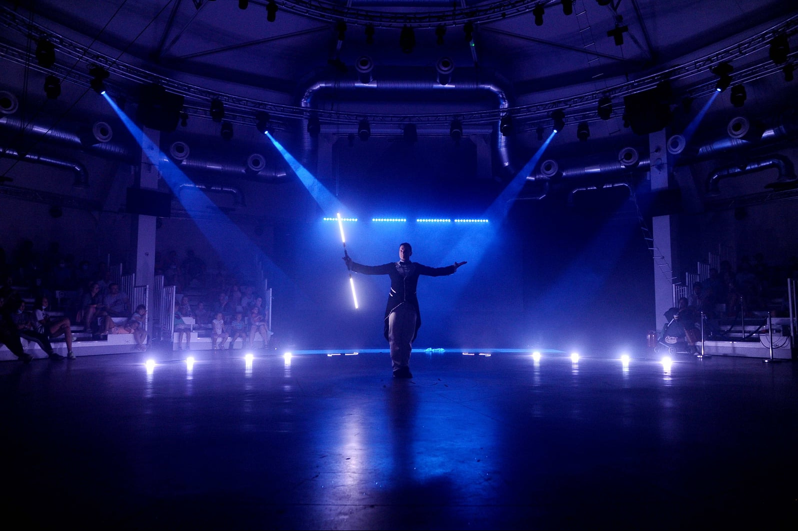 Oświetlona scena z osobą prezentującą lightshow z użyciem świetlnych rekwizytów, widzowie w tle, niebieskie światło, widowisko.