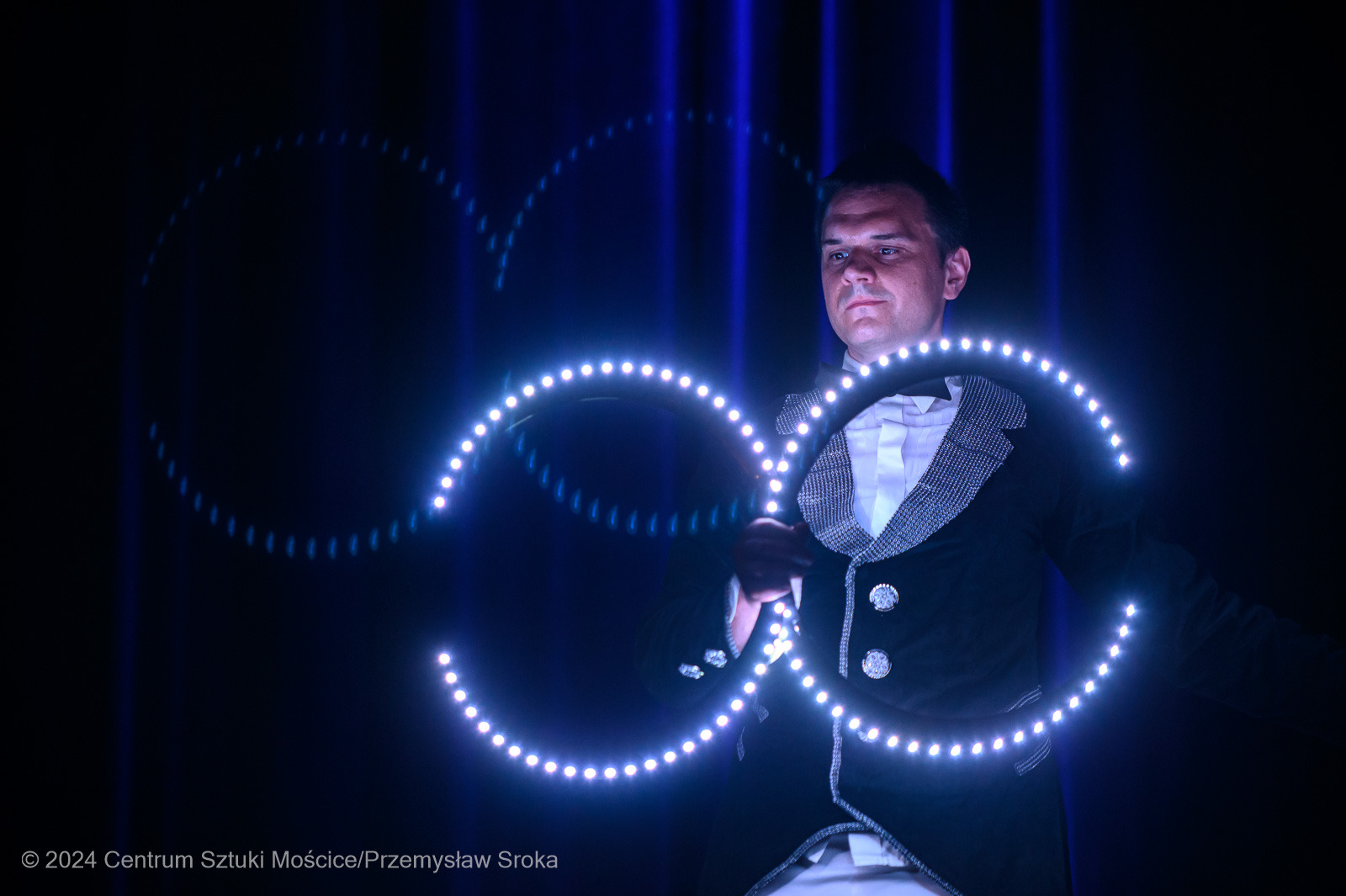 Mężczyzna w smokingu prezentuje lightshow z użyciem rekwizytów LED, tworząc świetlne wzory na ciemnym tle. Widoczne niebieskie pionowe pasy światła.