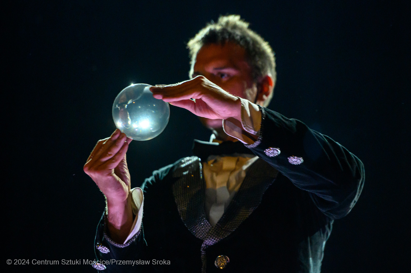 Artysta w czarnym fraku żongluje szklaną kulą na ciemnym tle, tworząc iluzję lewitacji. Oświetlenie podkreśla refleksy na kuli i detale stroju.