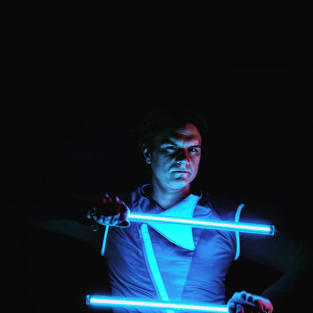 Mężczyzna w niebieskim oświetleniu trzymający świetlne pałeczki. Ubrany w kostium z elementami LED, na ciemnym tle. Wyraziste oświetlenie twarzy.