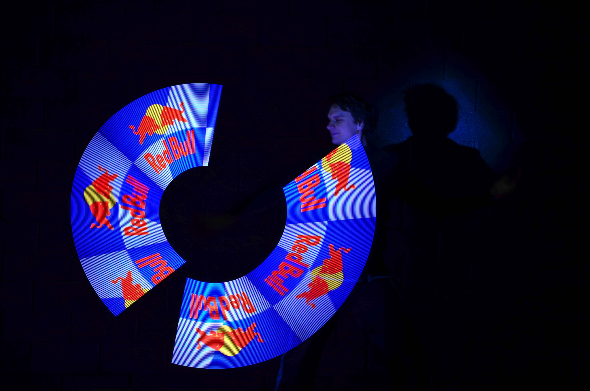 Mężczyzna wykonuje pokaz świetlny z logotypem Red Bulla w ciemności, tworząc dynamiczne efekty wizualne. Widoczny cień postaci i fragment ściany.