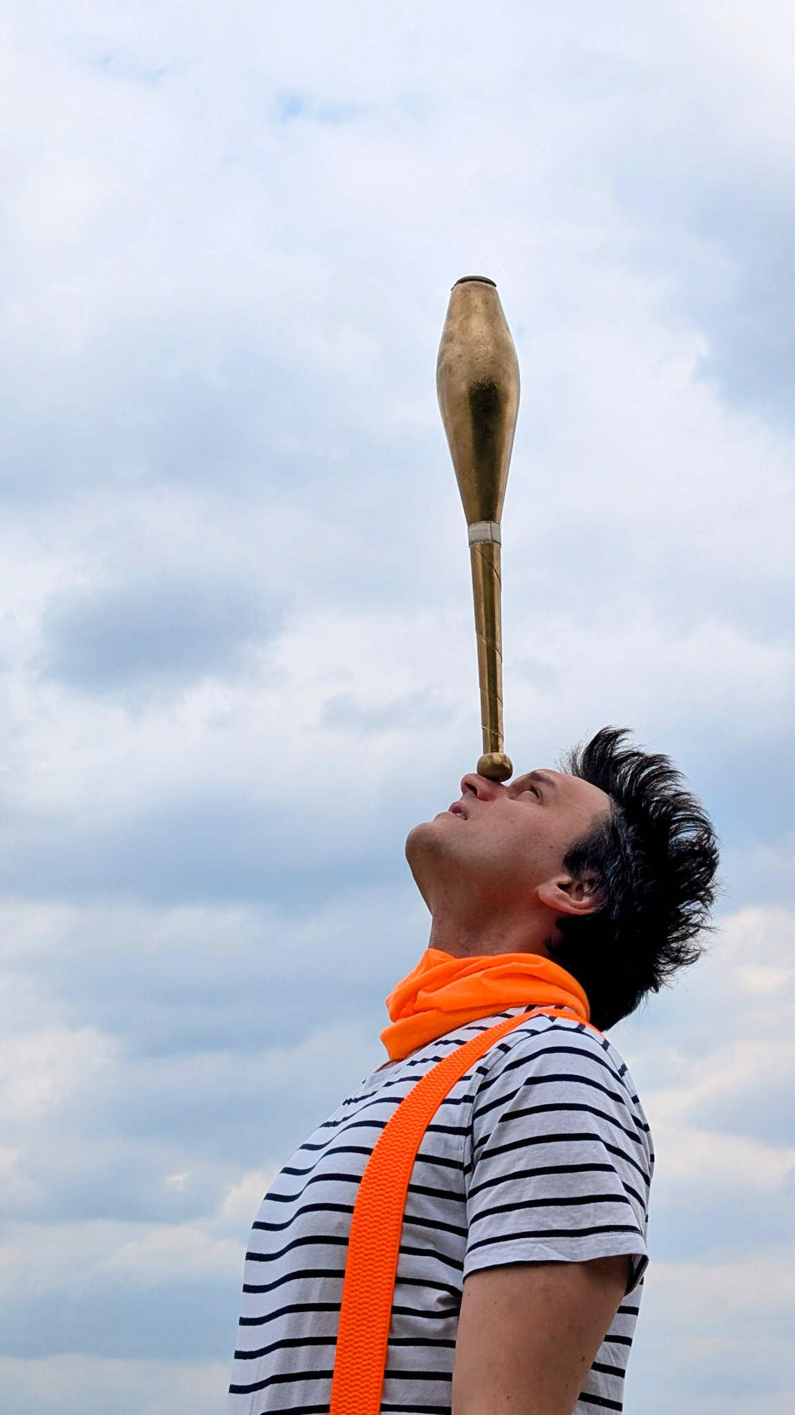 Artysta cyrkowy balansuje maczugą na nosie, patrząc w górę, na tle pochmurnego nieba. Ubrany w pasiastą koszulkę i pomarańczowy szal.