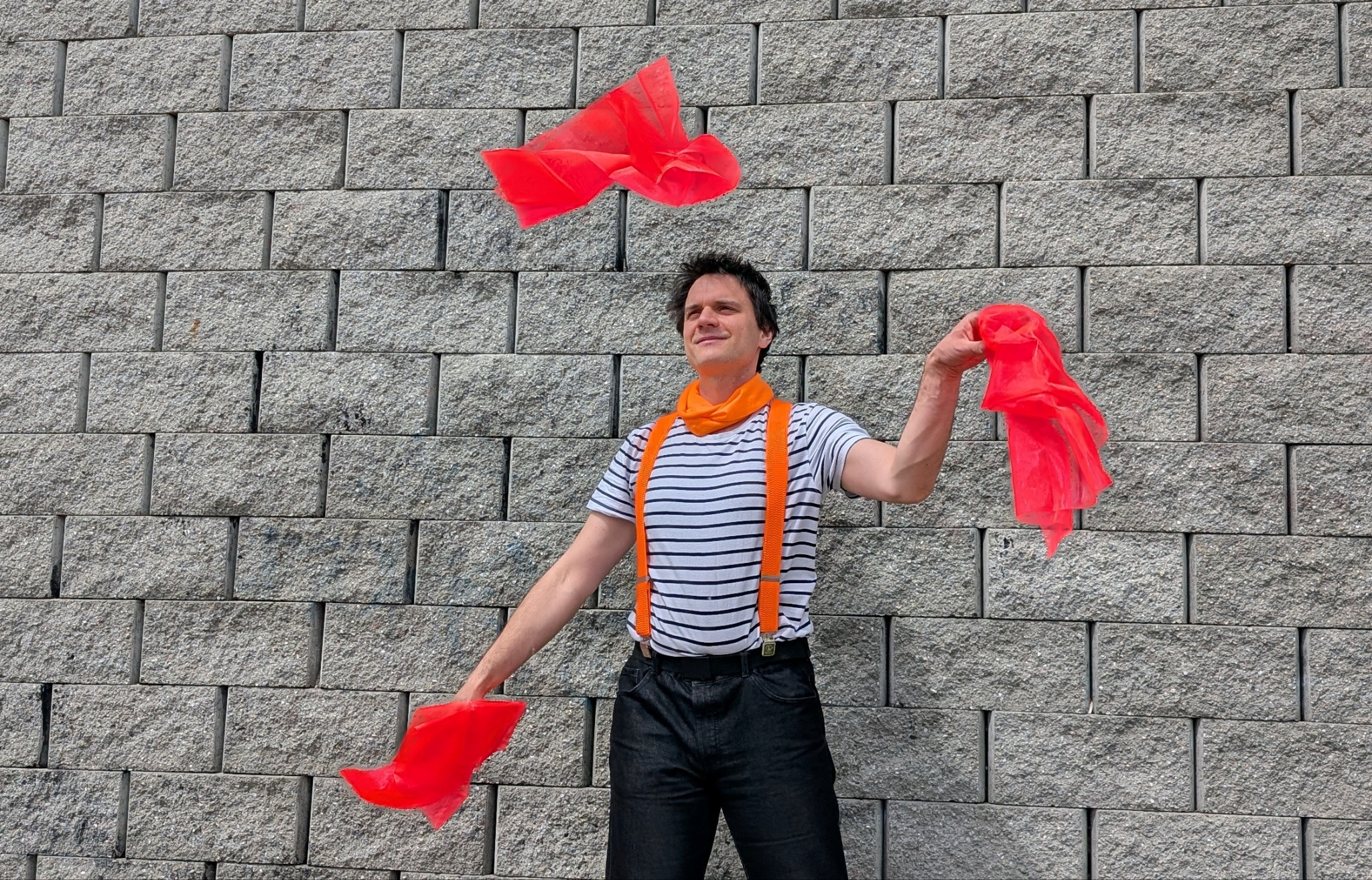 Żongler w pasiastym stroju i pomarańczowych szelkach, żonglujący trzema czerwonymi chustami na tle szarej ściany z bloków.