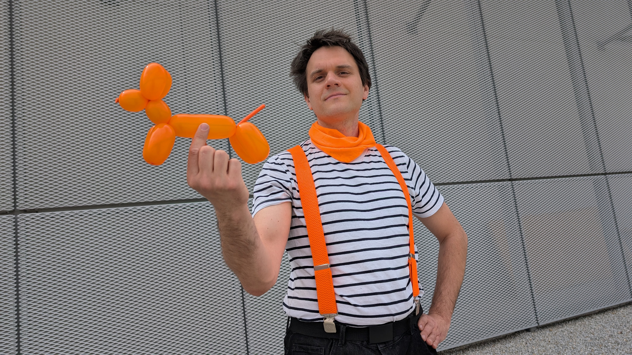 Animator w pasiastym stroju prezentuje pomarańczowego psa z balonów na tle metalowej ściany. Uśmiechnięty artysta z szalem i szelkami. Cyrkowa atmosfera.