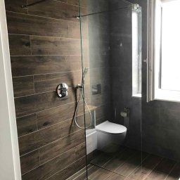 Dawid andraczek - Nowoczesna łazienka z kabiną prysznicową typu walk-in, wykończona płytkami imitującymi drewno i szarą glazurą. Widoczne chromowane elementy armatury i biała toaleta.