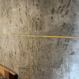 Dawid andraczek - Dekoracyjna ściana z efektem betonu i złotymi listwami w Nakle Śląskim. Fragment wezgłowia łóżka w dolnym rogu kadru. Nowoczesny design wnętrza.