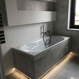 Dawid andraczek - Nowoczesna łazienka z wanną obudowaną szarą płytką, podświetloną listwą LED. Na ścianie półki wnękowe, okno z czarną ramą. Minimalistyczny design.