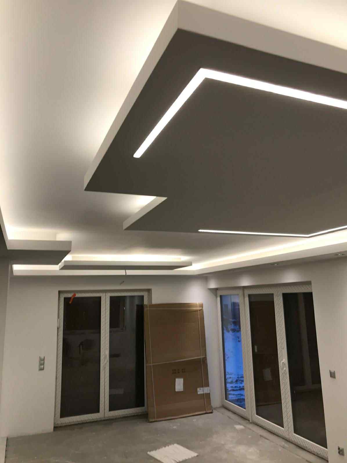 Nowoczesny sufit podwieszany z oświetleniem LED w kształcie geometrycznym, szare wnęki, białe obramowanie, widok na okna i kartonowe pudła na podłodze.