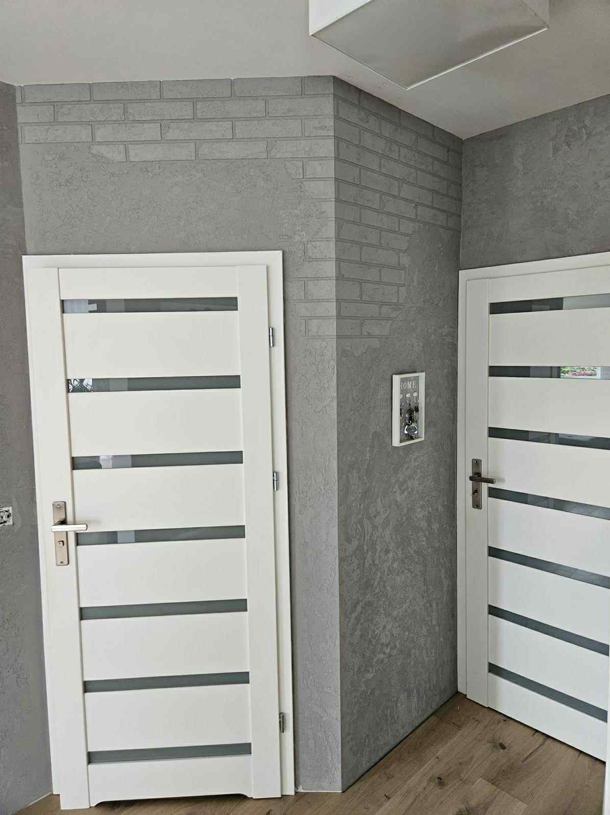 Szara ściana z efektem cegły i dekoracyjną fakturą, dwie białe drzwi z przeszkleniami, drewniana podłoga. Nowoczesne wykończenie wnętrza.