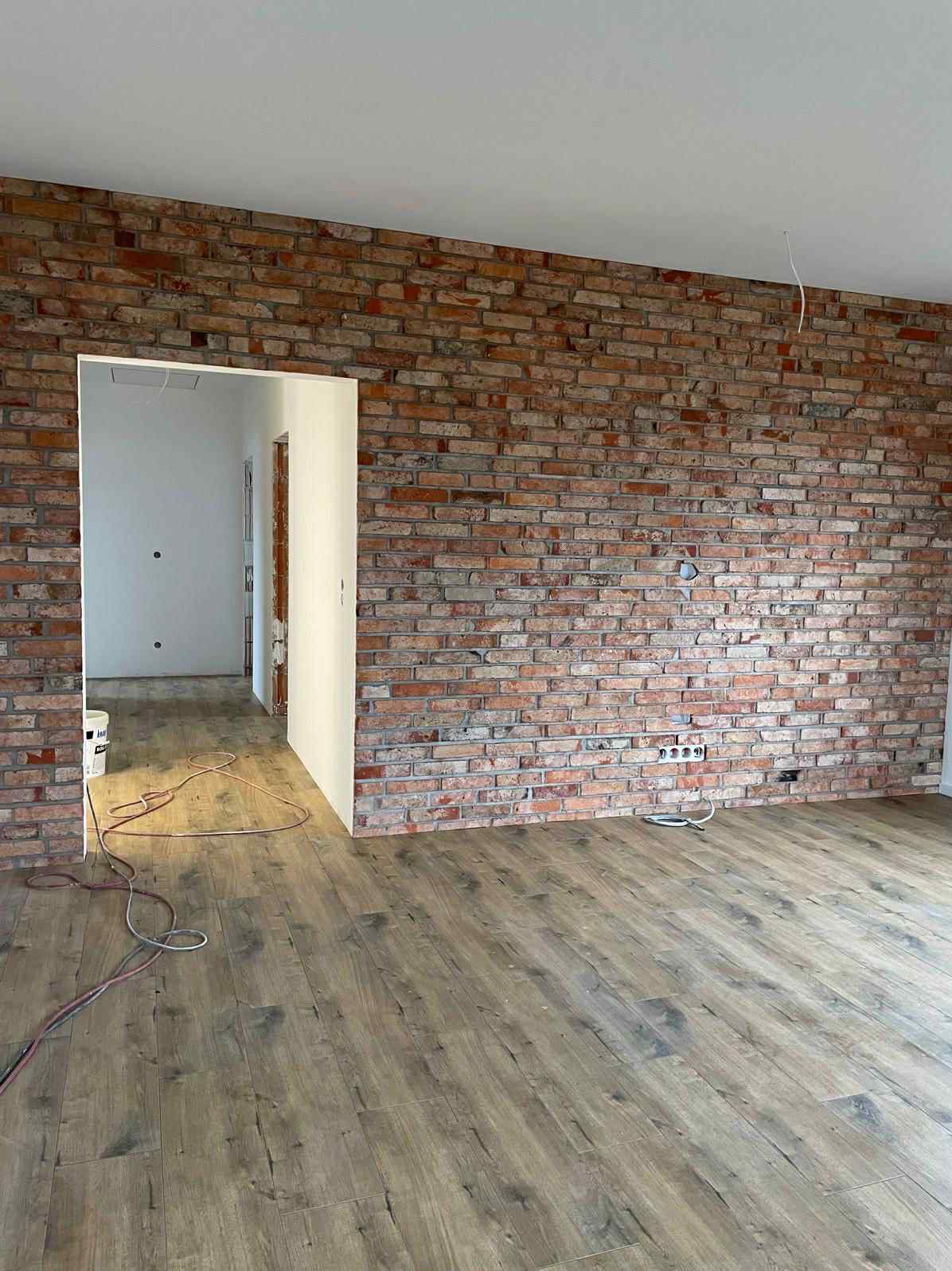 Ściana z cegły dekoracyjnej w nowym wnętrzu, z widocznymi gniazdkami elektrycznymi i drewnianą podłogą. W tle widoczne przejście do innego pomieszczenia.