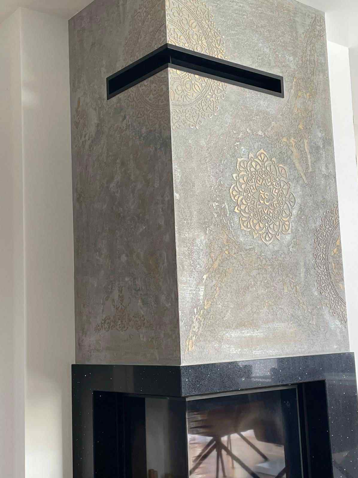 Ozdobny komin z szarego tynku strukturalnego z motywami mandali w odcieniach złota, wykończony czarnym kamieniem. Nowoczesny design.