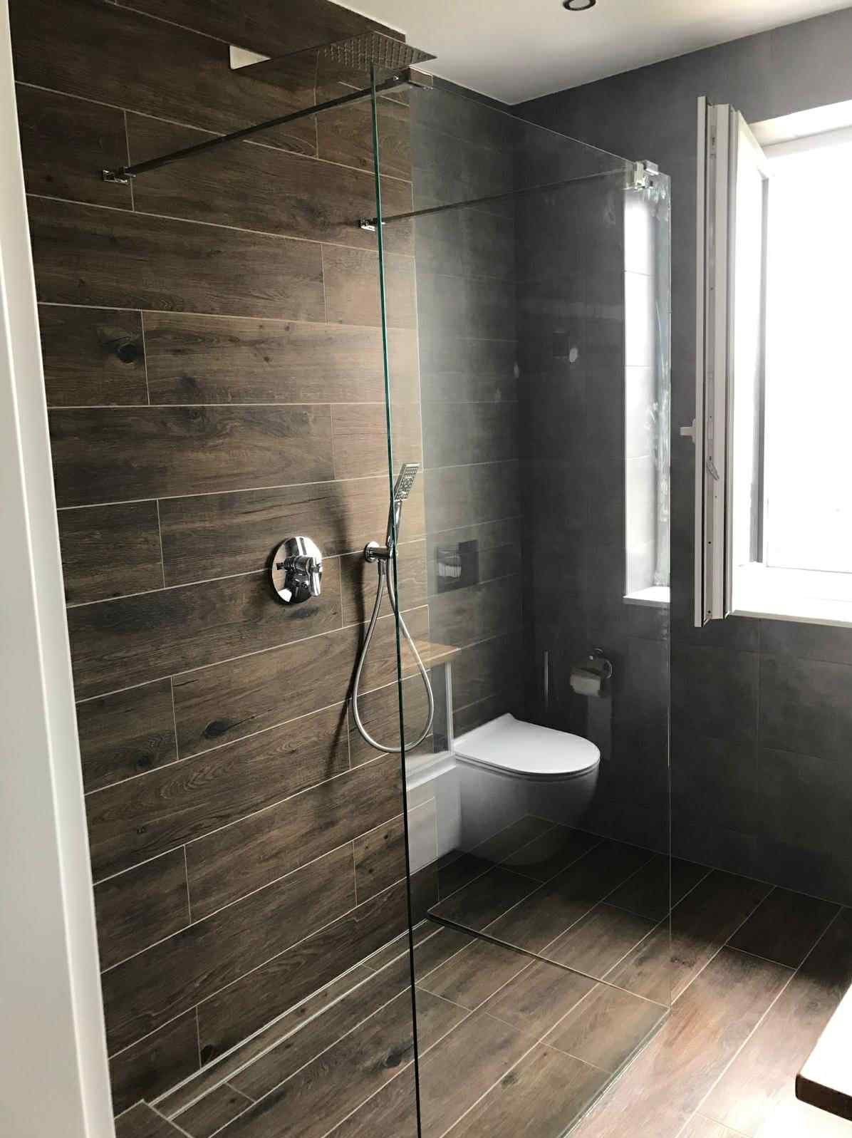 Nowoczesna łazienka z kabiną prysznicową typu walk-in, wykończona płytkami imitującymi drewno i szarą glazurą. Widoczne chromowane elementy armatury i biała toaleta.