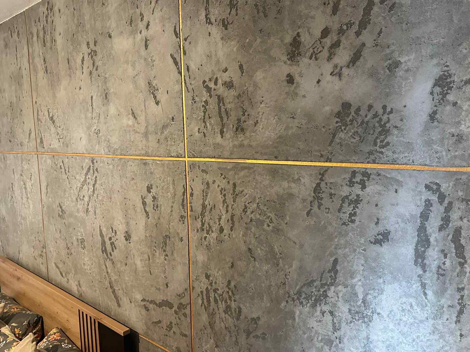 Dekoracyjna ściana z efektem betonu i złotymi listwami w Nakle Śląskim. Fragment wezgłowia łóżka w dolnym rogu kadru. Nowoczesny design wnętrza.
