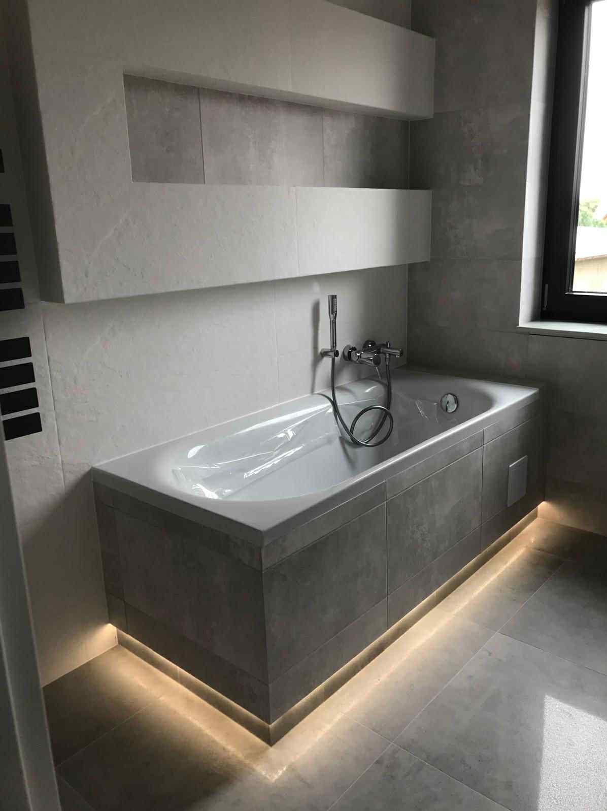 Nowoczesna łazienka z wanną obudowaną szarą płytką, podświetloną listwą LED. Na ścianie półki wnękowe, okno z czarną ramą. Minimalistyczny design.