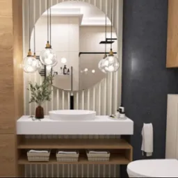 Nowoczesna łazienka z okrągłym lustrem, wiszącymi lampami, białą umywalką i drewnianymi szafkami. Minimalistyczny design z rośliną w wazonie.