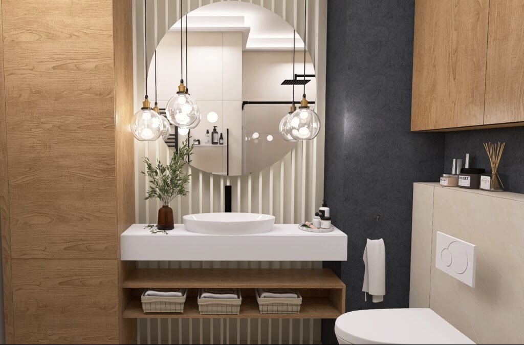 Nowoczesna łazienka z okrągłym lustrem, wiszącymi lampami, białą umywalką i drewnianymi szafkami. Minimalistyczny design z rośliną w wazonie.