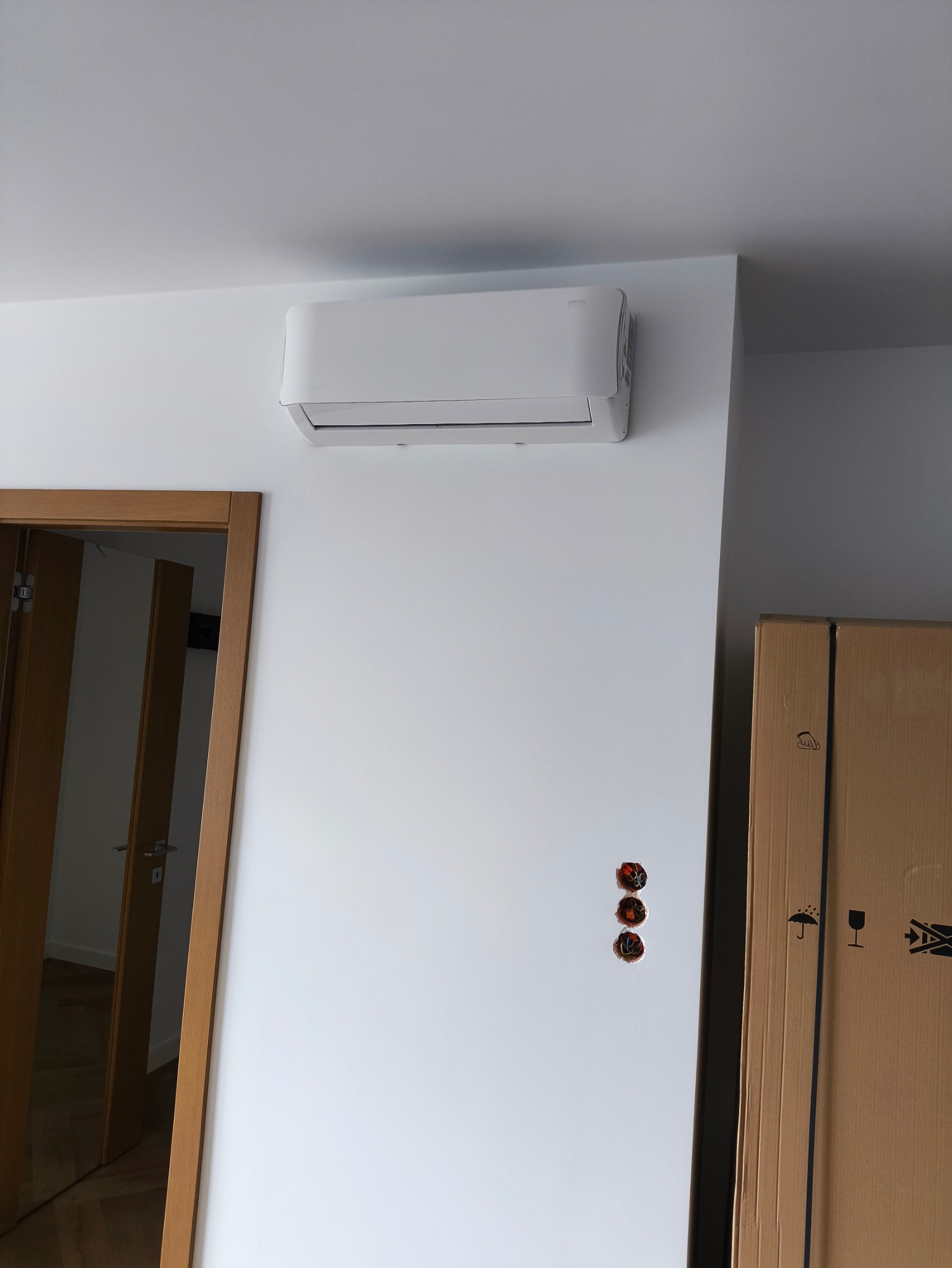 Biały klimatyzator ścienny w jasnym wnętrzu, widoczne przygotowane otwory na instalację elektryczną i kartonowe pudło. Minimalistyczny design.