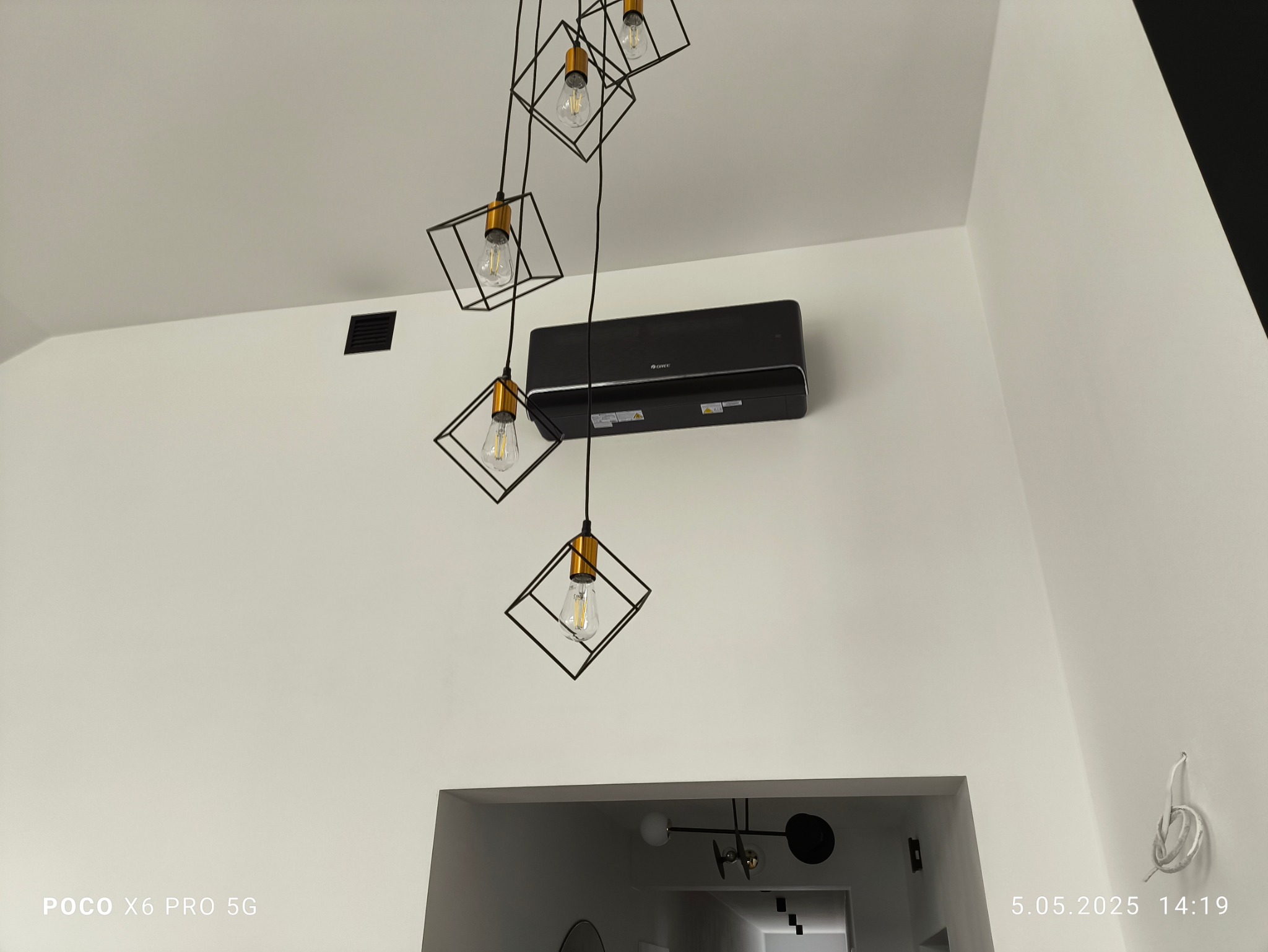 Czarny klimatyzator Gree zamontowany na białej ścianie w nowoczesnym wnętrzu, oświetlony przez minimalistyczną lampę z żarówkami w geometrycznych oprawach.