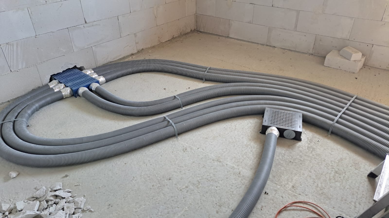 Szare, elastyczne rury wentylacyjne ułożone na podłodze w surowym wnętrzu, prowadzące do niebieskiej skrzynki rozdzielczej w ścianie z betonu komórkowego.