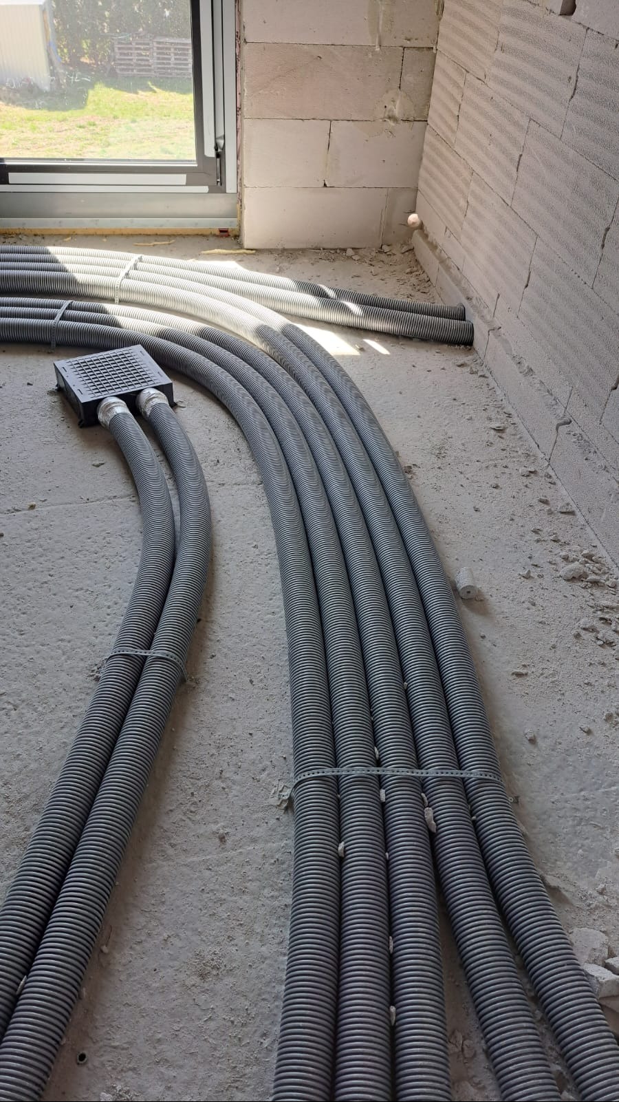 Szare, karbowane rury wentylacyjne ułożone równolegle na surowej podłodze w nowym budynku, z oknem w tle i widoczną instalacją podtynkową.