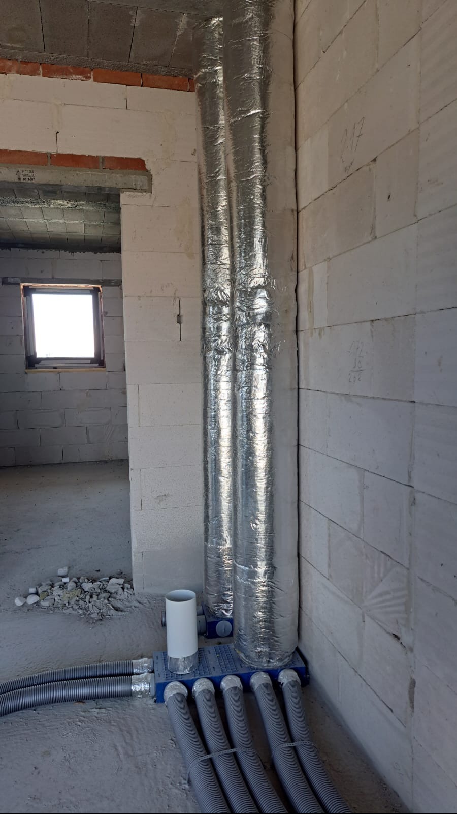 Dwie pionowe rury wentylacyjne w srebrnej izolacji, podłączone do niebieskiej skrzynki rozdzielczej z szarymi przewodami w surowym wnętrzu budynku.