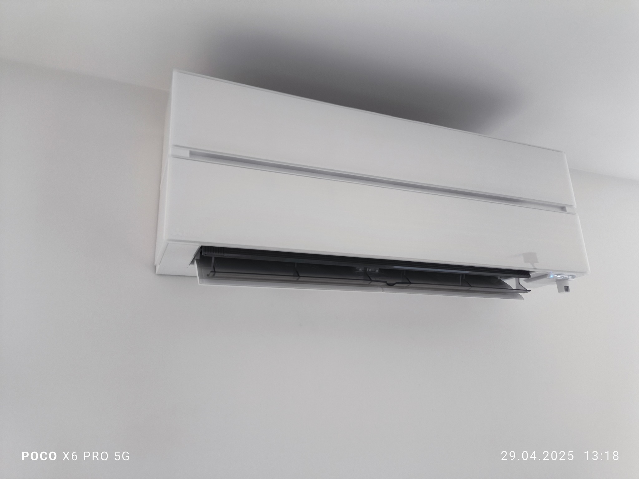 Biała klimatyzacja ścienna zamontowana w jasnym pomieszczeniu. Widoczny detal w postaci otwartej klapy wylotowej powietrza. Minimalistyczny design.