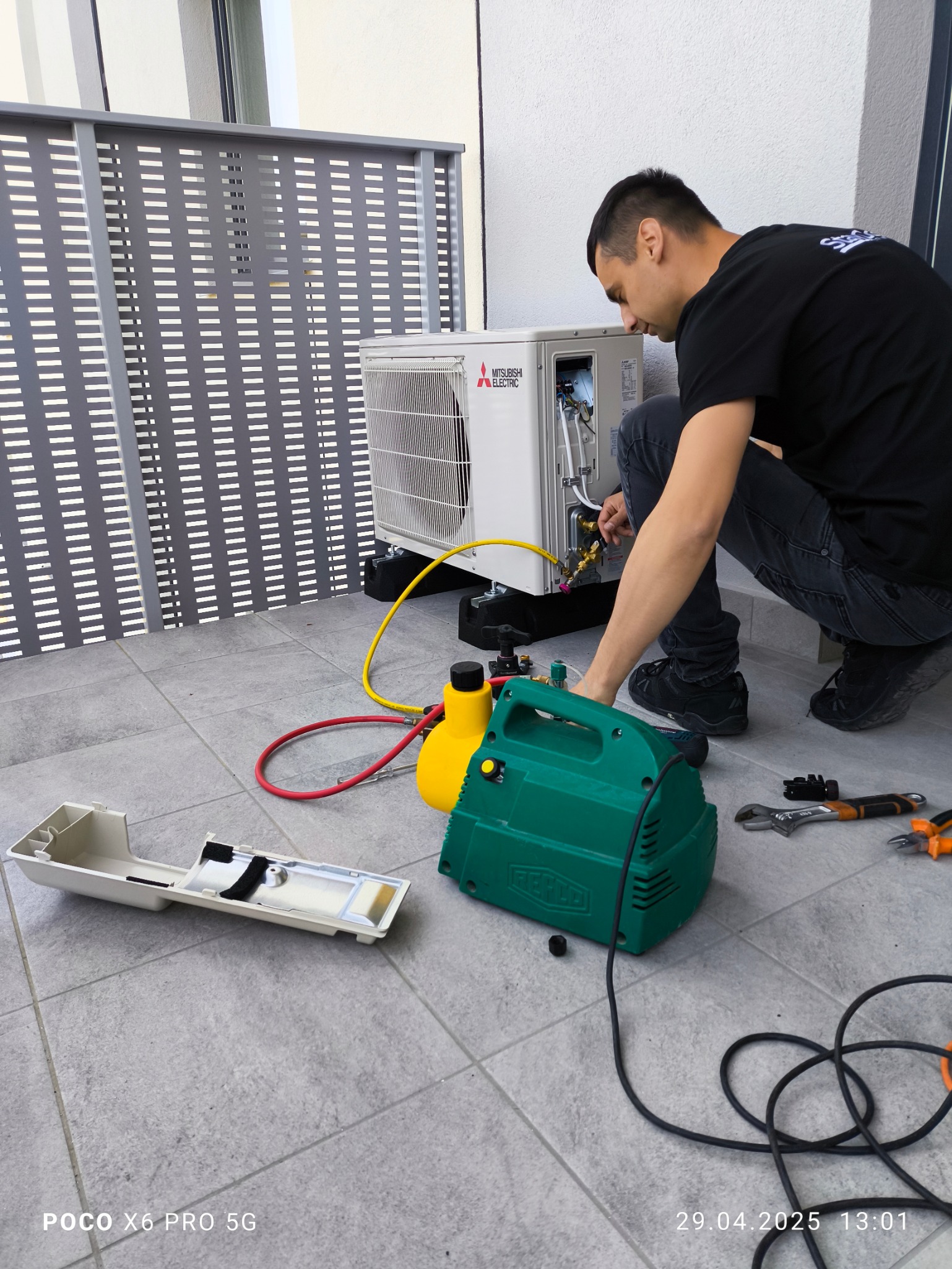 Serwisant w trakcie montażu klimatyzacji Mitsubishi Electric na balkonie, używając specjalistycznych narzędzi i pompy próżniowej. Widoczne przewody i elementy instalacji.