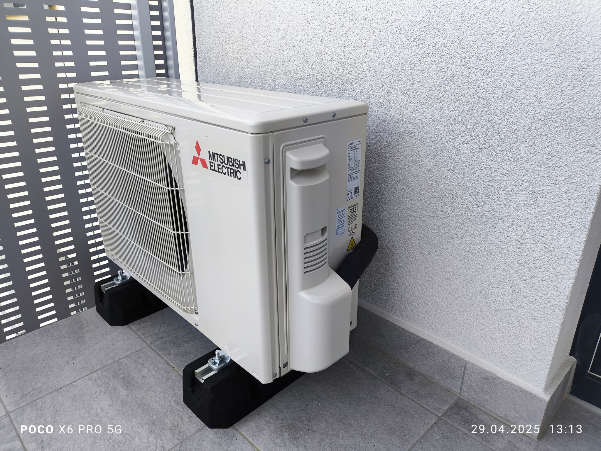 Zewnętrzna jednostka klimatyzacji Mitsubishi Electric na balkonie, zamontowana na czarnych podkładkach antywibracyjnych. Widoczna ażurowa balustrada i strukturalna ściana.