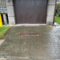 Mokry bruk po umyciu, widoczny odpływ liniowy. Na krawędzi żółty pojemnik, w tle brązowe drzwi garażowe i zieleń. Częściowo umyta kostka.