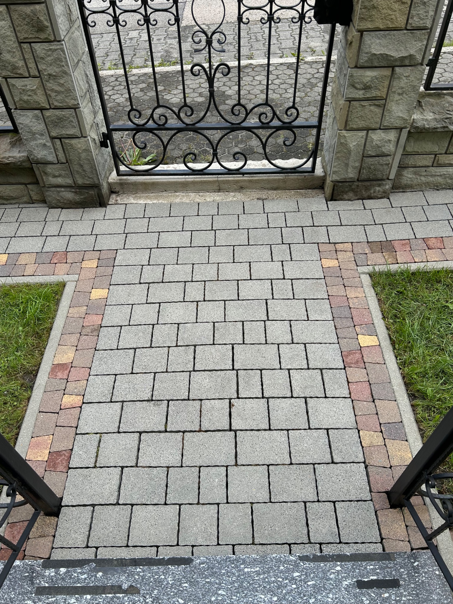 Ułożona kostka brukowa prowadząca do ozdobnej, metalowej bramy, z widocznymi fragmentami trawnika po bokach. Kąt ujęcia z góry podkreśla regularność wzoru.