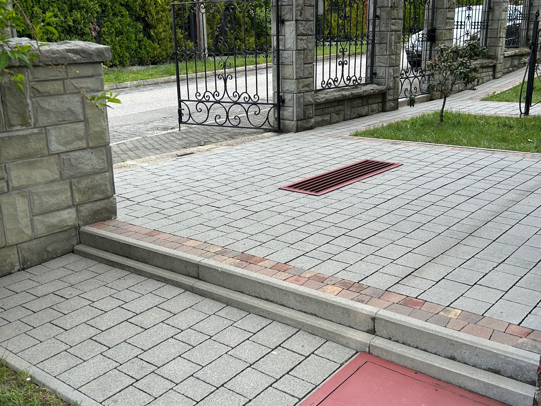 Szara kostka brukowa ułożona w jodełkę, z czerwoną kratką ściekową i obrzeżami z kostki w ciepłych barwach, prowadząca do ozdobnej, czarnej bramy.