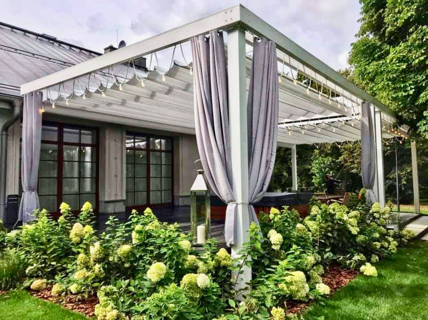 Elegancka, biała pergola z szarymi zasłonami i dekoracyjnym oświetleniem, otoczona kwitnącymi hortensjami w ogrodzie. Widoczny fragment domu w tle.