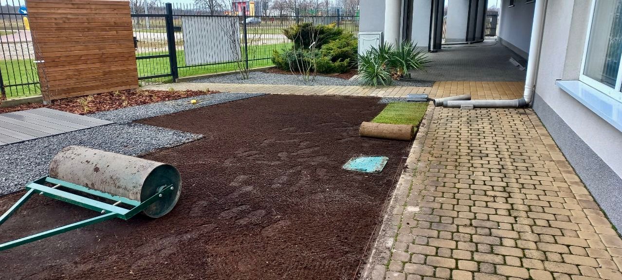 Przygotowany teren pod trawnik z rolki, częściowo rozłożona trawa, walec ogrodowy, kostka brukowa, elementy dekoracyjne z kamienia i drewna. Widok z góry.