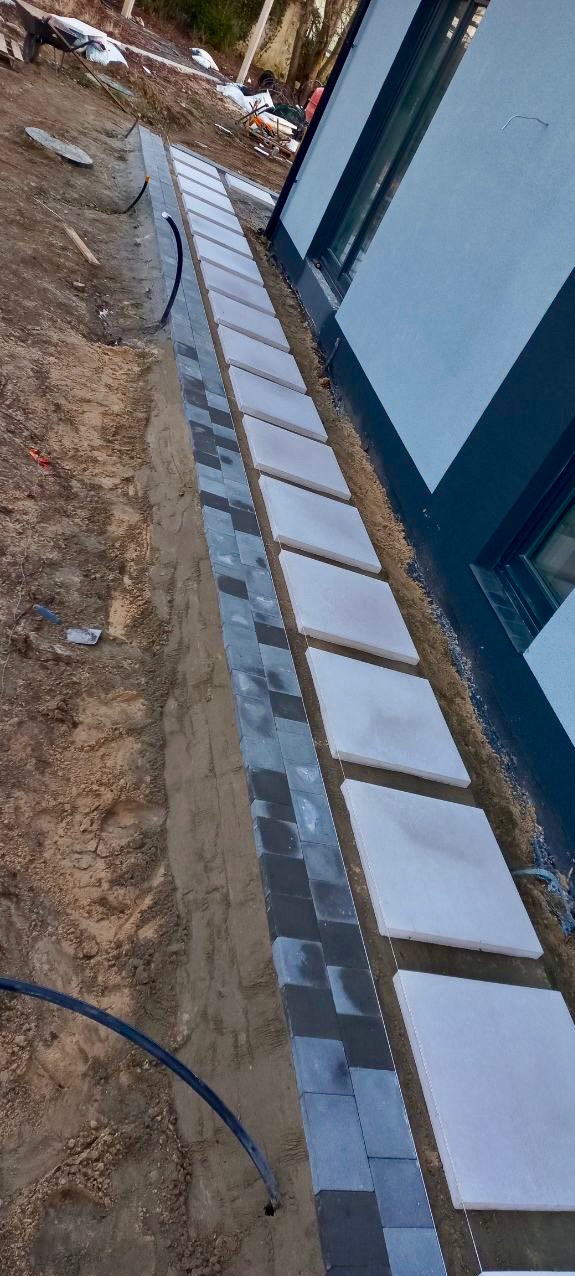 Układanie chodnika z szarych i białych płyt przy ścianie budynku. Widoczny wykop i czarna rura. Prace w toku, etap budowy ścieżki.