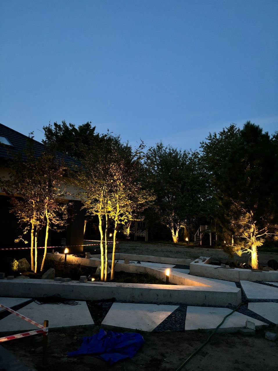 Oświetlony ogród po zmroku z betonowymi elementami i młodymi drzewami podświetlonymi lampami. Widoczne elementy instalacji elektrycznej i taśma ostrzegawcza.