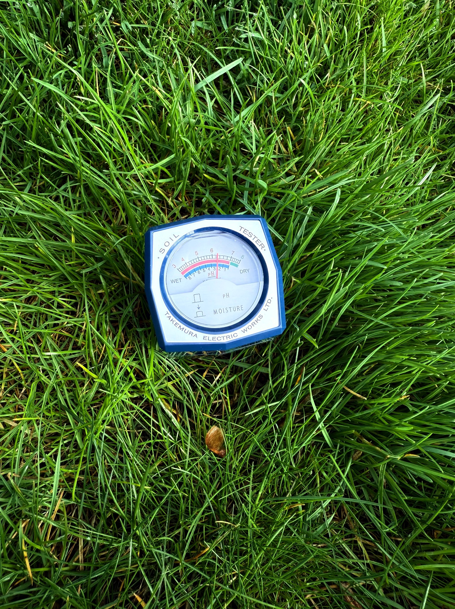 Miernik pH gleby Takemura wbity w gęstą, zieloną trawę. Urządzenie wskazuje odczyn lekko kwaśny. Pomiar kwasowości gleby w trawniku.