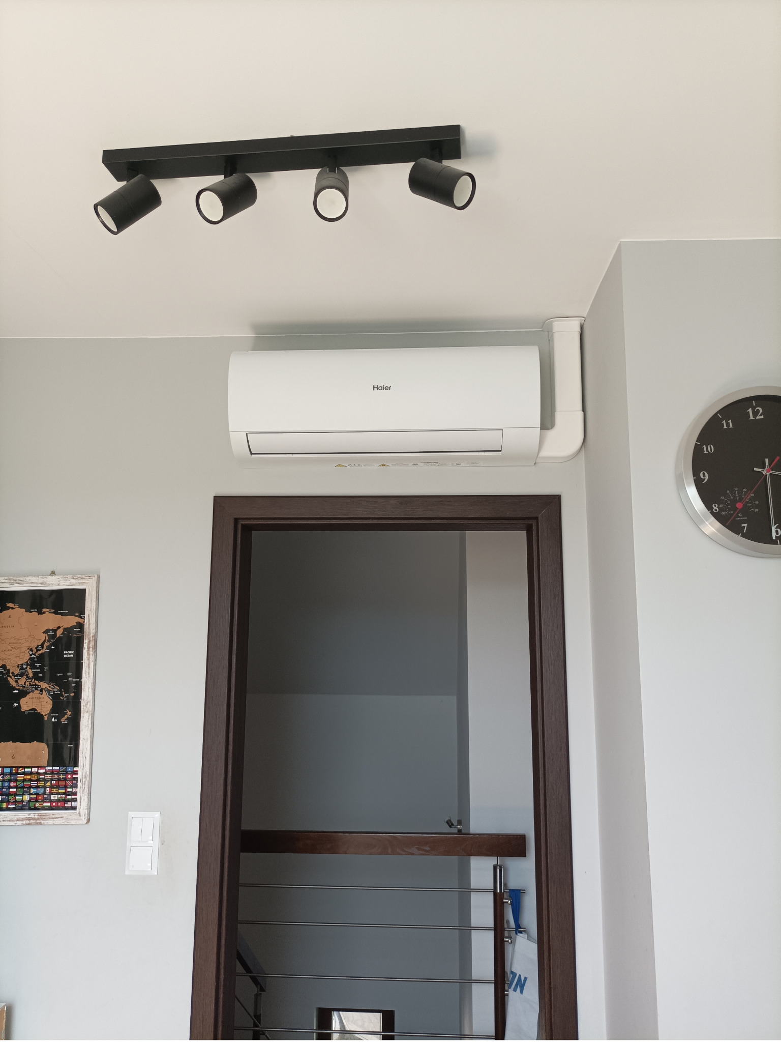 Biały klimatyzator Haier zamontowany nad brązowymi drzwiami w szarym pomieszczeniu z zegarem i mapą świata. Czarna listwa z reflektorami sufitowymi.