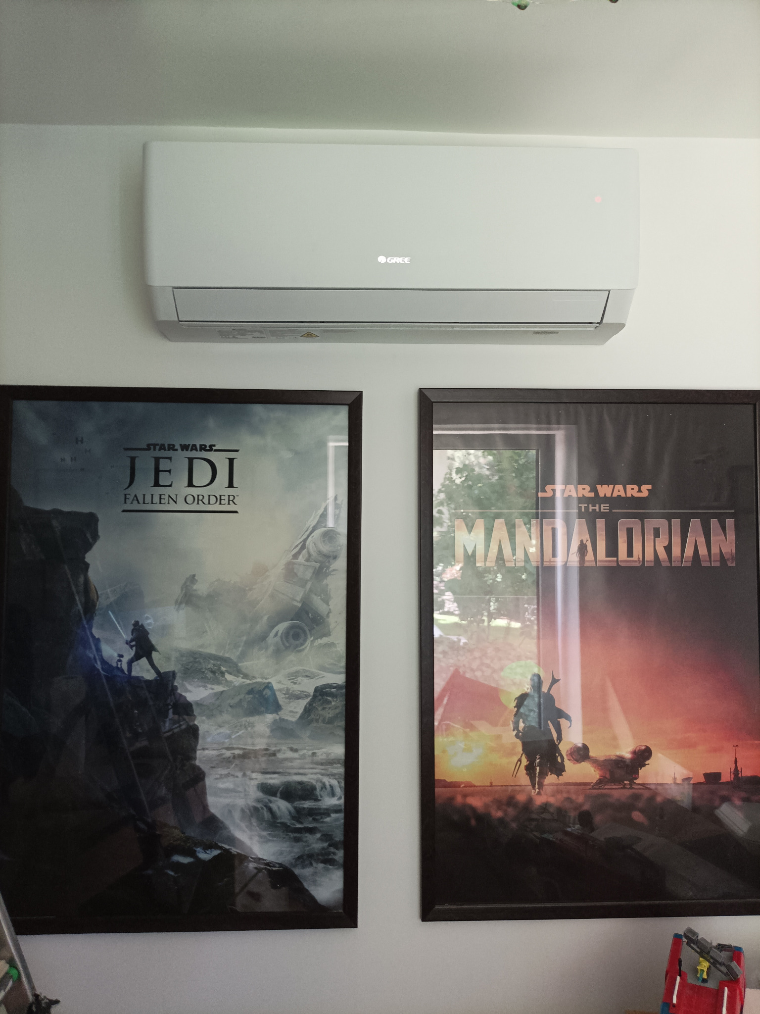 Biała klimatyzacja marki Gree zamontowana na ścianie, pod nią dwa plakaty Star Wars: Jedi Fallen Order i Mandalorian, tworząc nowoczesną aranżację wnętrza.